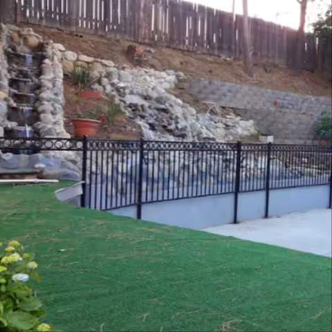 Landscaped outdoor area with artificial turf, a black metal fence, rock waterfall features and a wooden privacy fence.