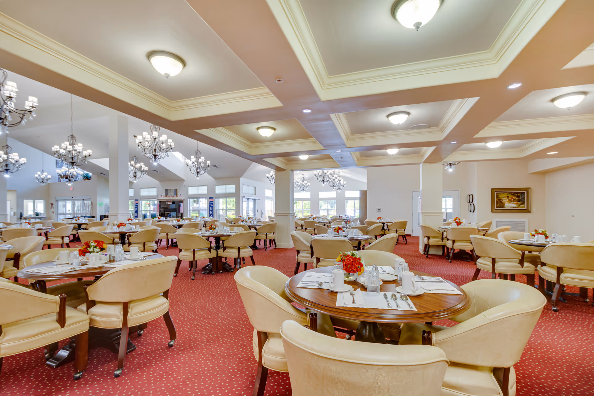 A spacious dining room in a senior living facility with multiple round wooden tables set with white cups, silverware, and small floral centerpieces. The room features beige upholstered chairs, red carpet with a subtle pattern, chandeliers, and large windows allowing natural light to fill the space.