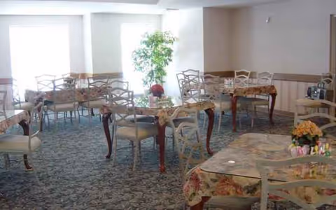 Bright senior living dining room with floral tablecloths, chairs, and potted plants.