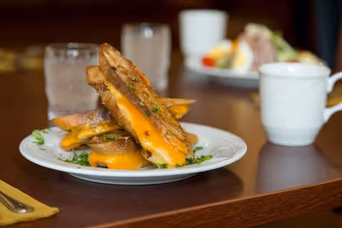 Grilled cheese sandwiches stacked on a white plate on a dining table with a cup and salad in the background.