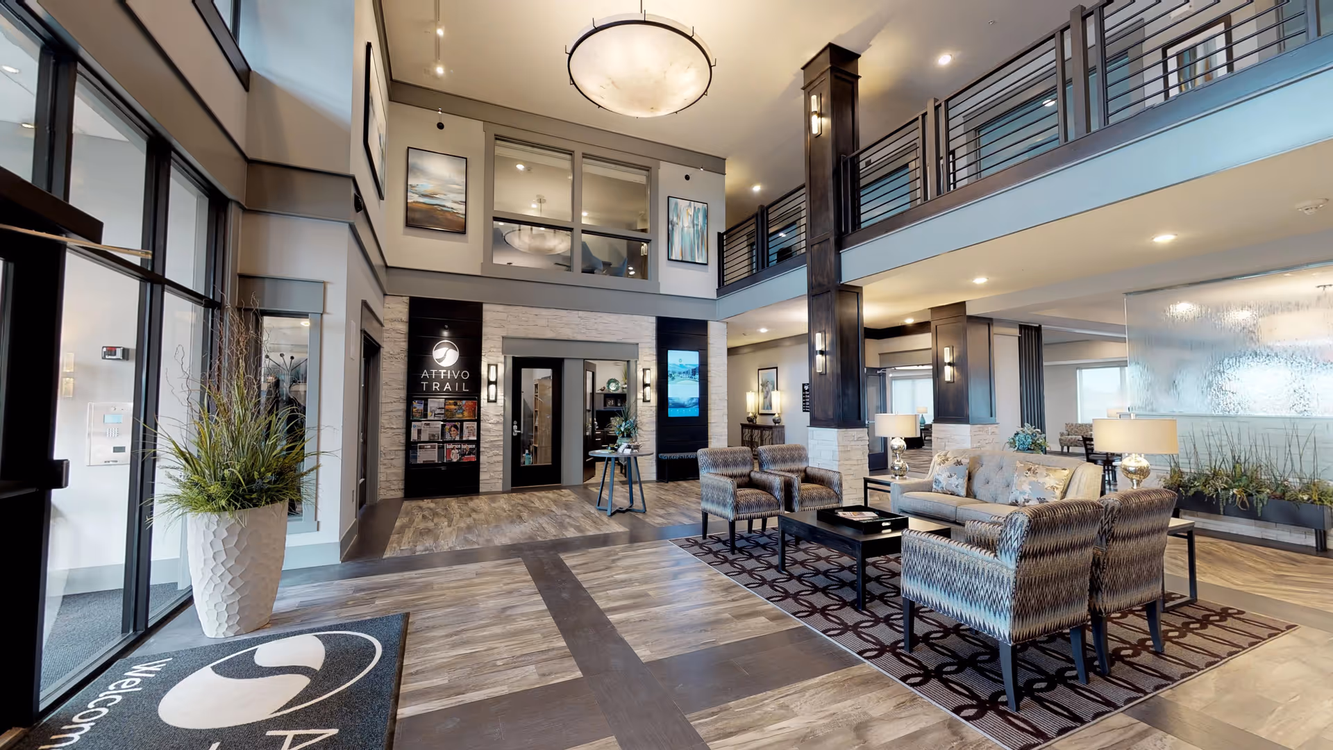 Spacious and modern senior living facility lobby with high ceilings, large windows, and a seating area including a sofa and chairs on a patterned rug. The entrance area features a welcome mat with the Attivo Trail logo, a tall plant in a white textured pot, and a wall with the Attivo Trail name and logo. The space is well-lit with ceiling lights and wall sconces, and has a second-floor balcony overlooking the lobby.