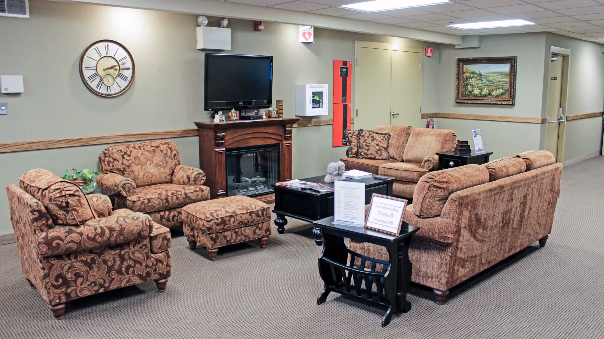 A cozy senior living facility common area with patterned armchairs, a matching ottoman, and two brown sofas arranged around a black coffee table and a small side table. A flat-screen TV is mounted above a wooden fireplace mantel. The walls are light green with a large clock and a framed landscape painting. The carpet is beige, and there are doors and hallway visible in the background.