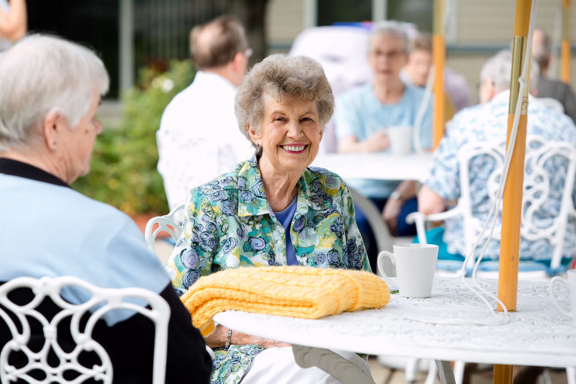 A group of elderly people sitting outdoors at white metal tables with umbrellas. One elderly woman in a colorful jacket is smiling at the camera, with a yellow knitted blanket and a white mug on the table in front of her. Other seniors are engaged in conversation in the background.