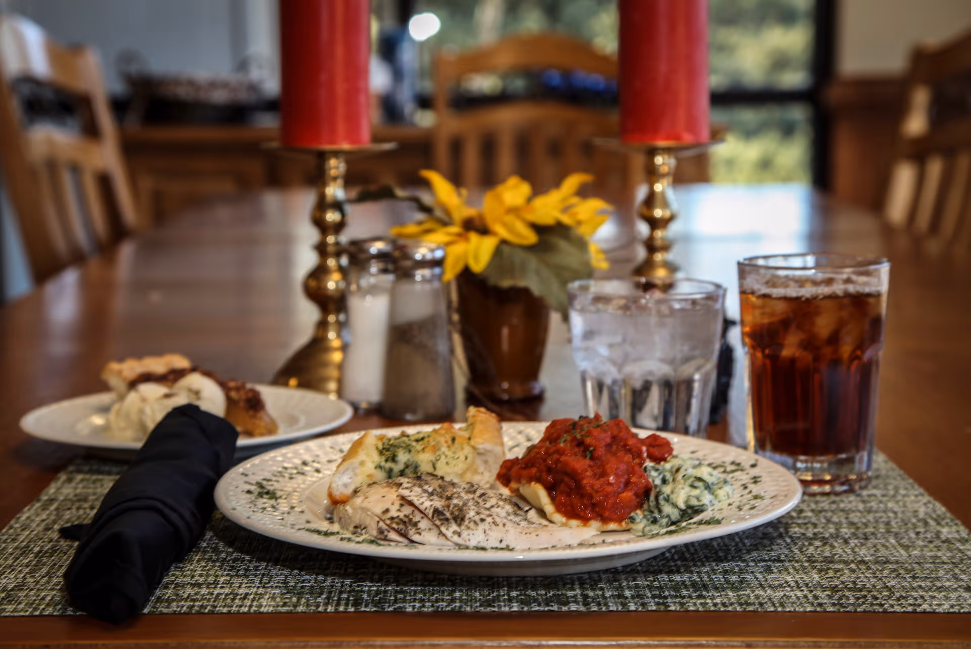 A dining table set with a plate of food including chicken with white sauce, ravioli with red sauce, and a side of spinach. There is a glass of iced tea, a glass of water, a black cloth napkin, two red candles in brass holders, salt and pepper shakers, and a small vase with yellow flowers on the table. In the background, wooden chairs and a window with greenery outside are visible.