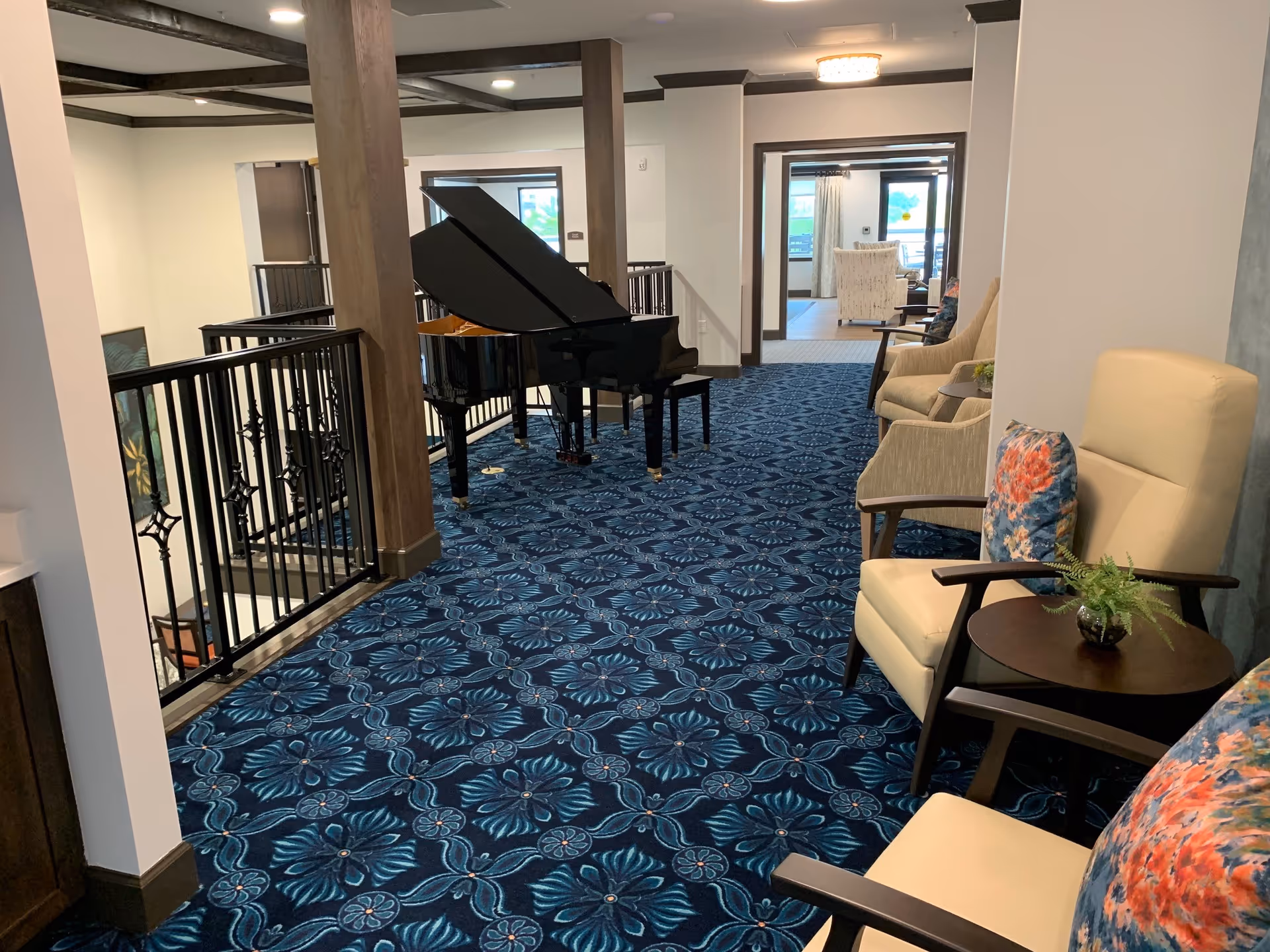 Interior hallway area with blue patterned carpet, featuring a black grand piano near a railing. On the right side, there are beige armchairs with colorful floral cushions and small round tables with plants. The hallway leads to another room with more seating and large windows.