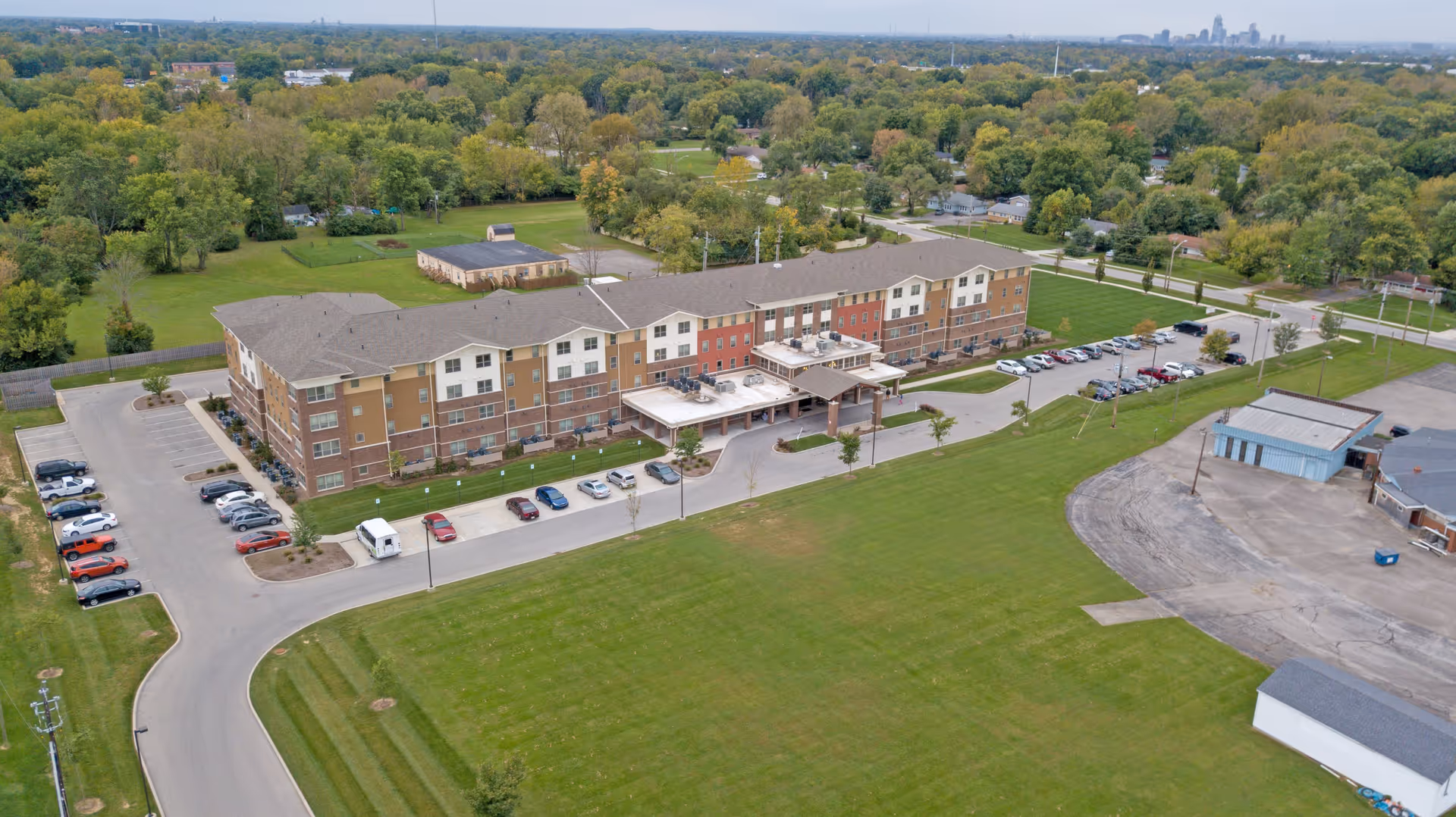 Aerial view of a large three-story senior living facility named Oasis At 30th, surrounded by green lawns, parking lots with cars, and trees in the background under a clear sky.