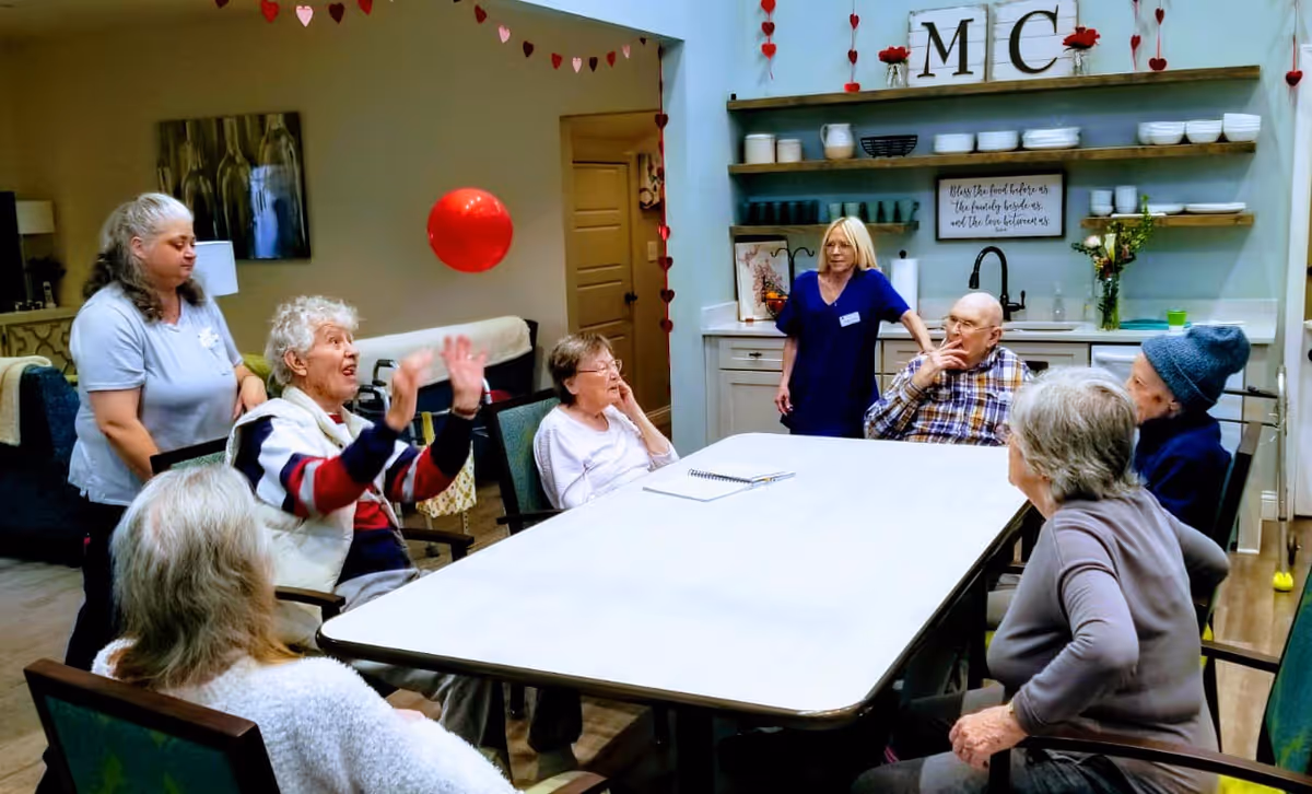 A group of elderly people and two caregivers gathered around a large rectangular table in a well-lit room with kitchen shelves and decorations in the background. One elderly woman is gesturing with her hands while others listen attentively. The room has heart-shaped decorations hanging on the walls.