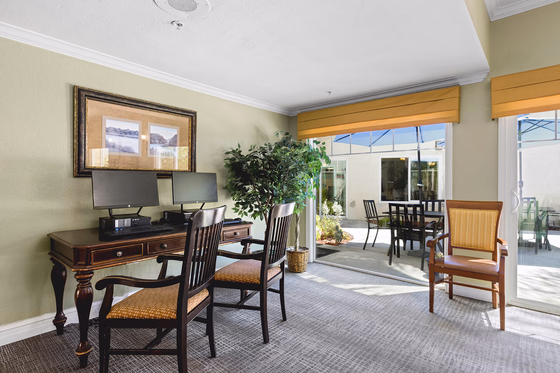 A bright lounge with a wooden desk holding two computer monitors, chairs, a potted plant, and sliding glass doors opening to an outdoor patio with tables.