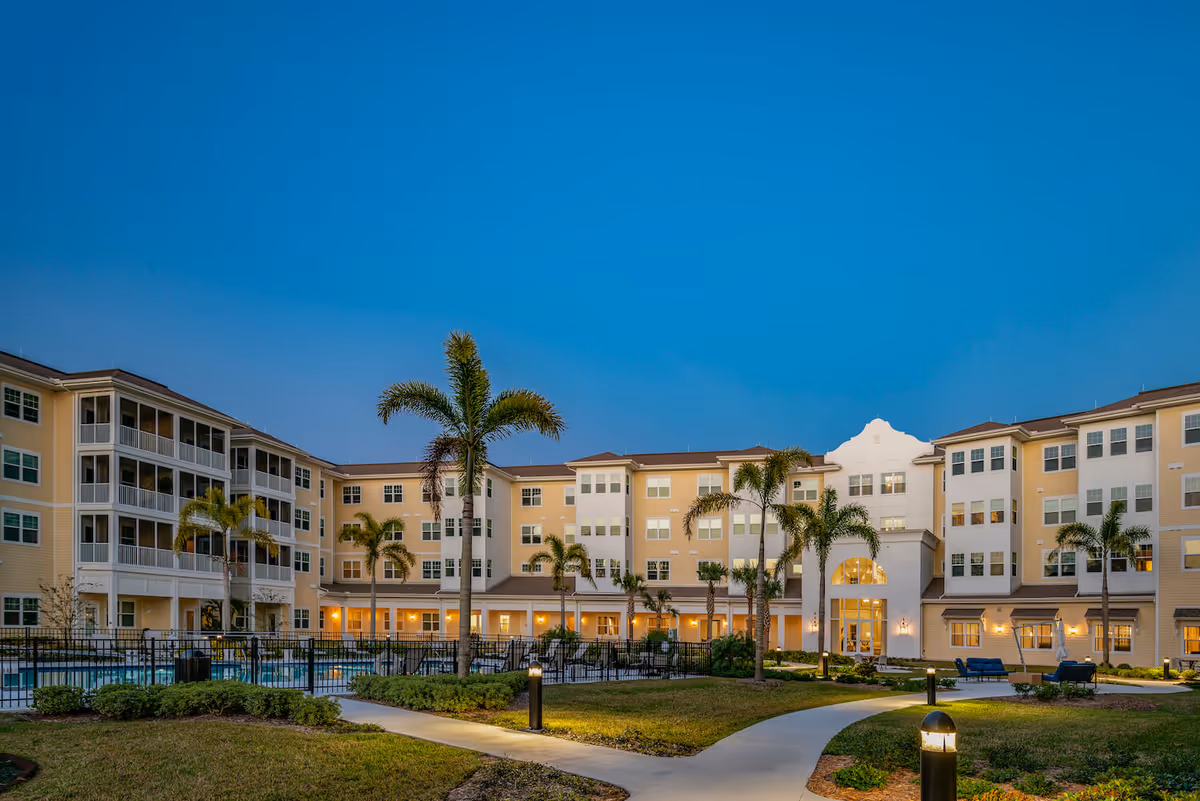 Exterior view of Arbor Terrace Citrus Park senior living facility at dusk, showing a large multi-story building with balconies, palm trees, a swimming pool area, and well-lit pathways surrounded by landscaped greenery.