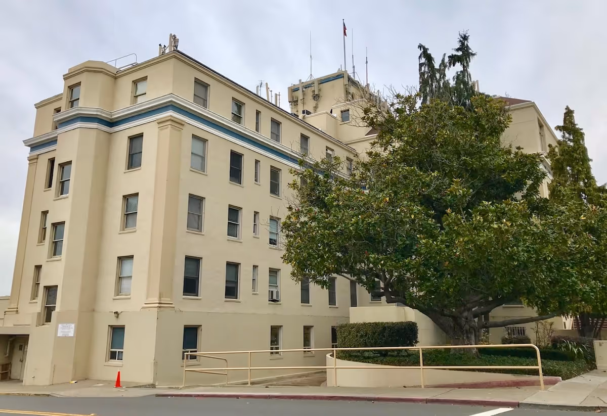 Exterior view of a multi-story beige hospital building with windows, a large tree in front, and a sidewalk ramp.