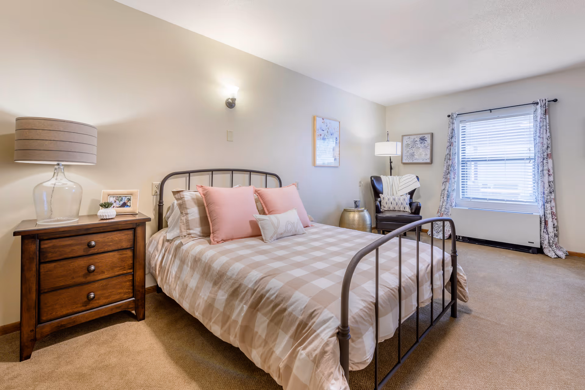 A cozy bedroom with a metal frame bed dressed in beige and white checkered bedding and pink pillows. To the left of the bed is a wooden nightstand with a glass lamp, a small plant, and a framed photo. On the right side of the room, there is a dark leather armchair with a white throw blanket and a patterned pillow, next to a round metallic side table and a floor lamp. The room has beige carpet, light-colored walls, a window with floral curtains, and framed artwork on the walls.