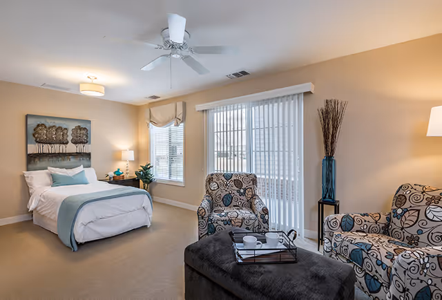 A cozy bedroom with a bed dressed in white linens and a light blue throw blanket. There is a nightstand with a lamp and decorative items next to the bed. Two patterned armchairs and a matching loveseat are arranged around a dark ottoman with a tray holding cups. The room has beige walls, a ceiling fan, a window with blinds, and sliding glass doors with vertical blinds leading outside.