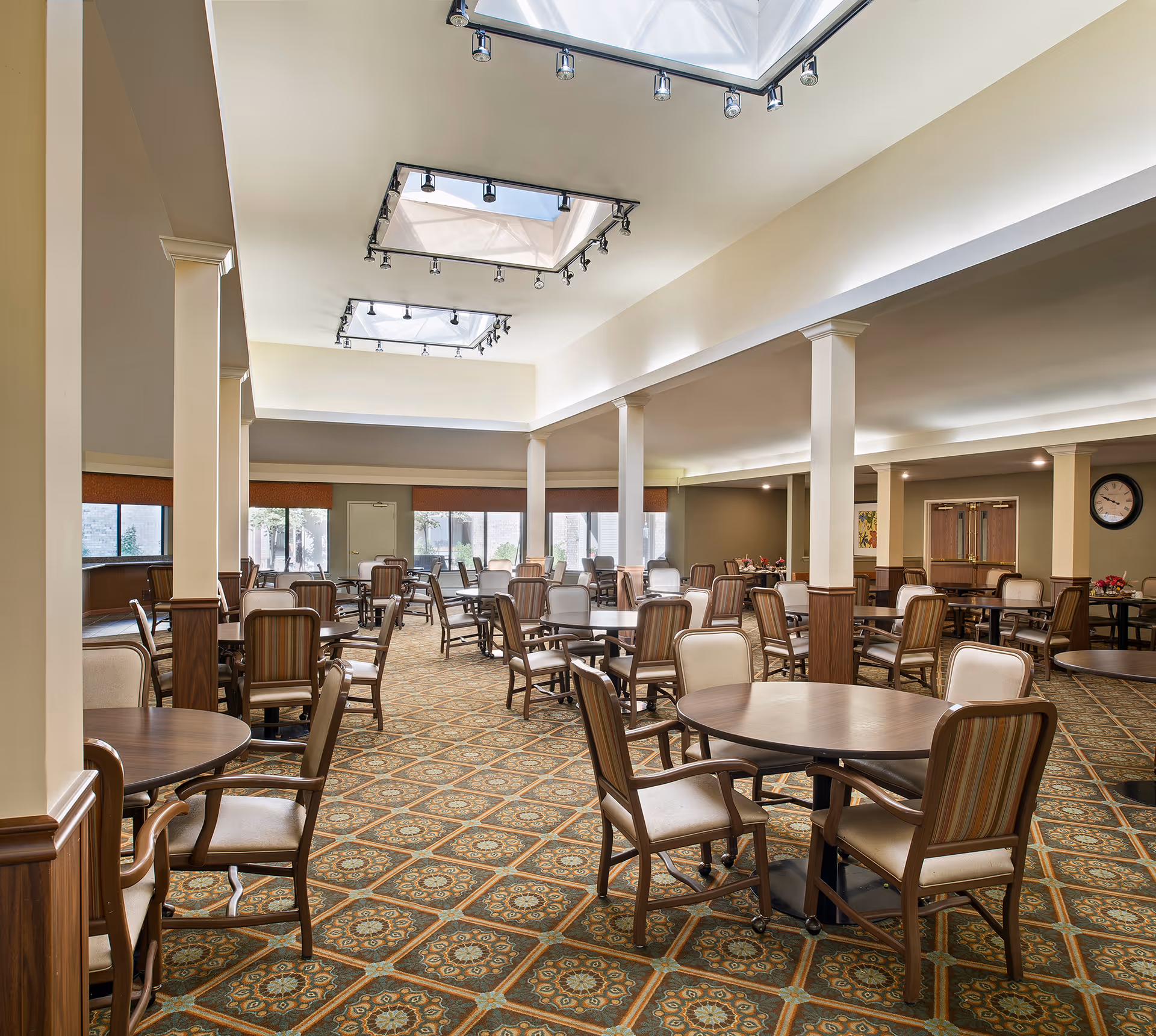 Spacious dining room with multiple round tables and chairs under skylights in a senior living facility.