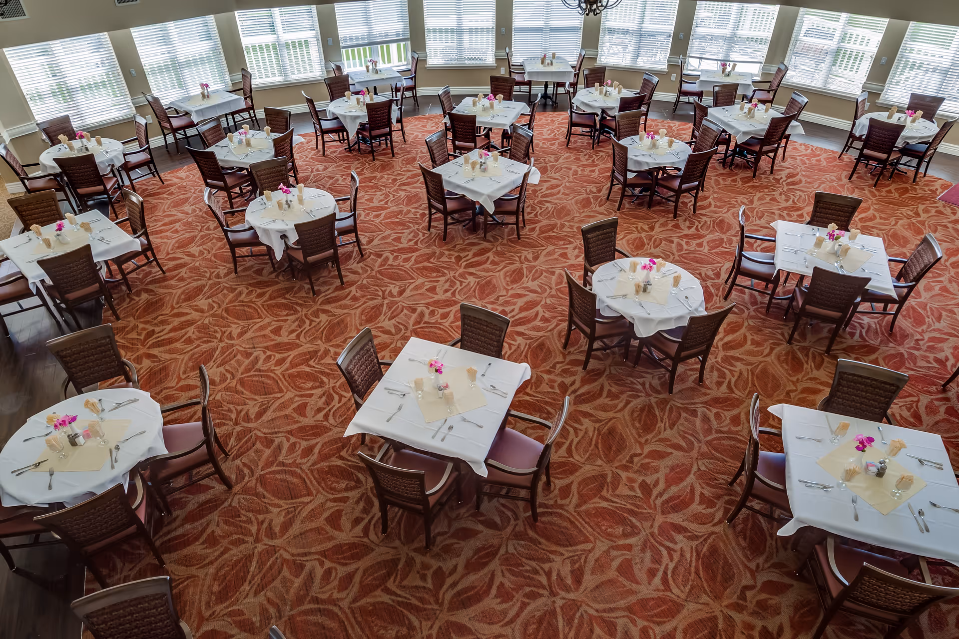 Spacious dining room with multiple round and square tables neatly set with white tablecloths and floral centerpieces on a patterned carpet under large windows.