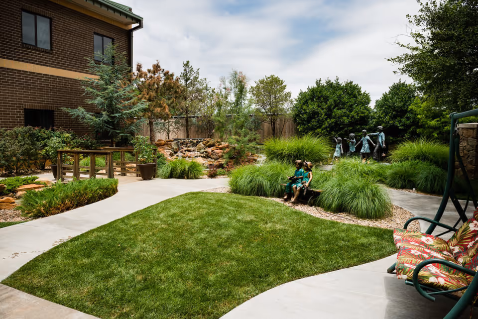 Manicured courtyard with green lawn, paved walkways, ornamental grasses, a small wooden bridge and bronze child statues beside a brick building.