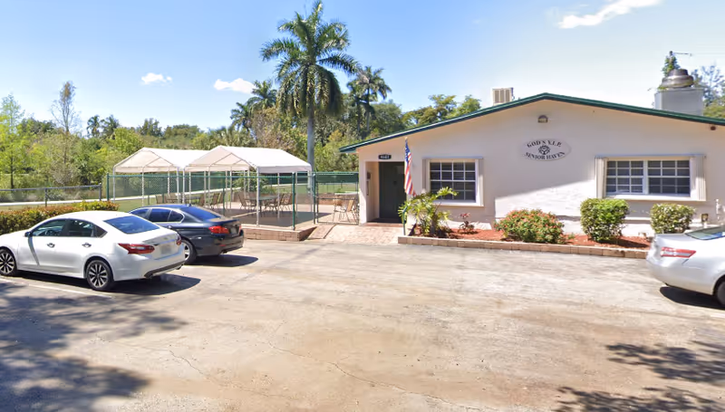 Exterior view of a single-story senior living facility building with a sign that reads 'God's VIP Senior Haven'. There are three cars parked in front of the building on a paved area. To the left of the building, there is a fenced outdoor area with white canopy tents. Palm trees and other greenery are visible in the background under a clear blue sky.