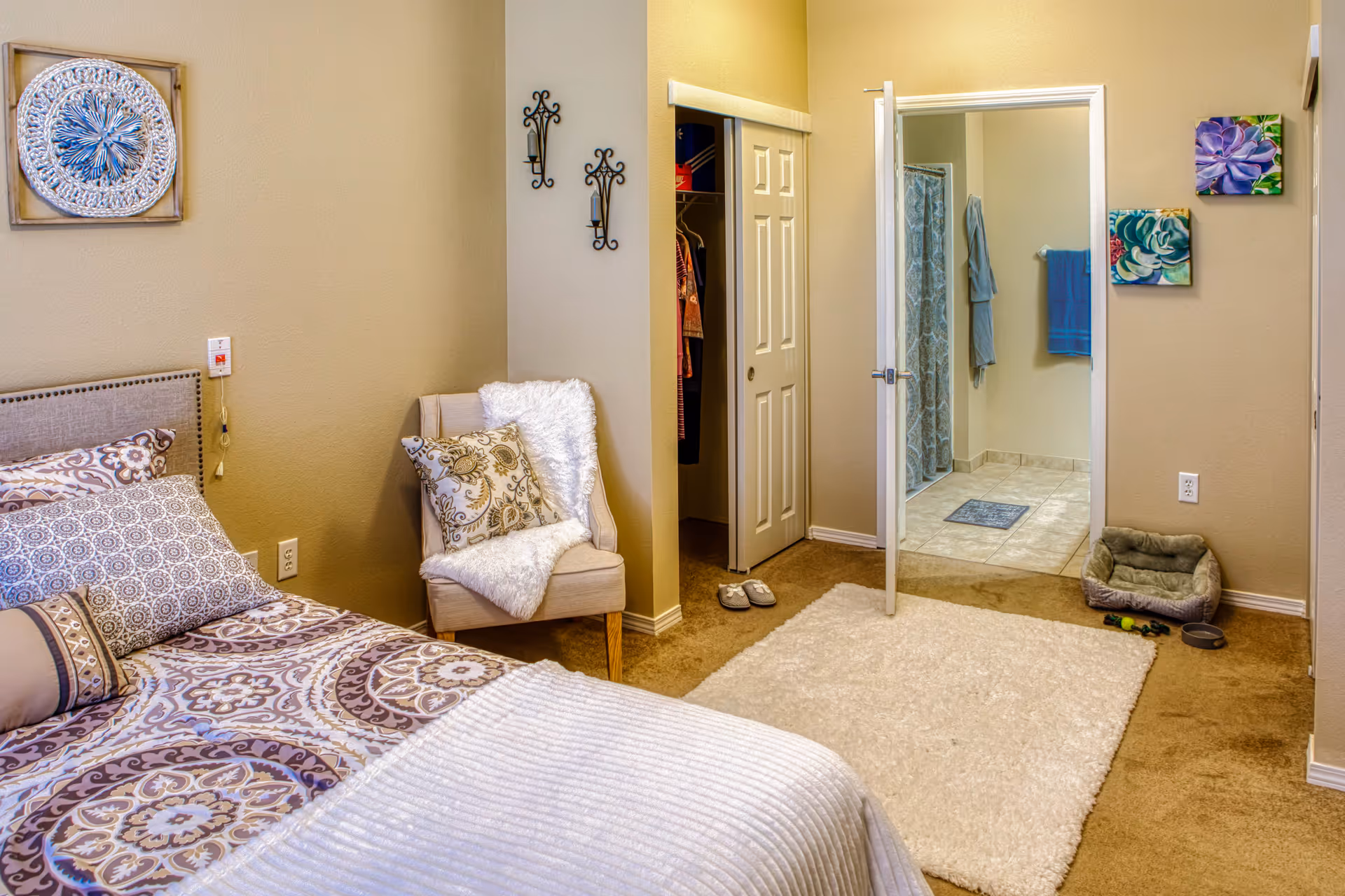 A cozy bedroom with a patterned bedspread and multiple pillows on the bed. There is a beige armchair with a decorative pillow and a white throw blanket next to the bed. The room has beige walls adorned with wall art and candle holders. An open door reveals a bathroom with tiled flooring, a shower curtain, and blue towels hanging on the wall. A small pet bed with toys and a water bowl is placed on the carpet near the bathroom door.