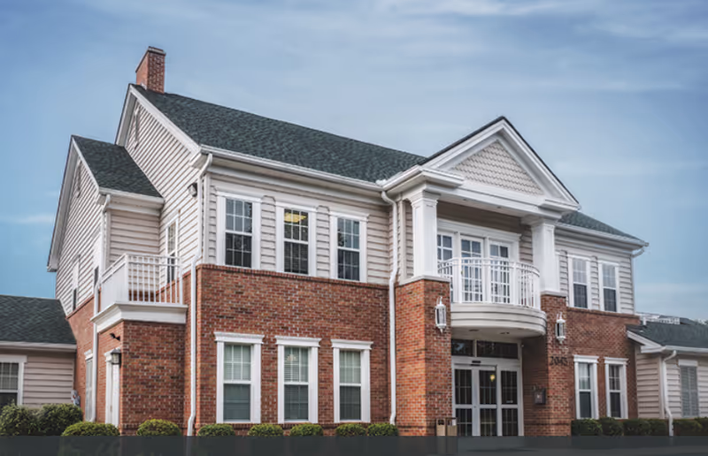 Exterior view of a two-story senior living facility building with a combination of red brick and beige siding, multiple windows, a small balcony above the main entrance, and a green roof under a clear sky.