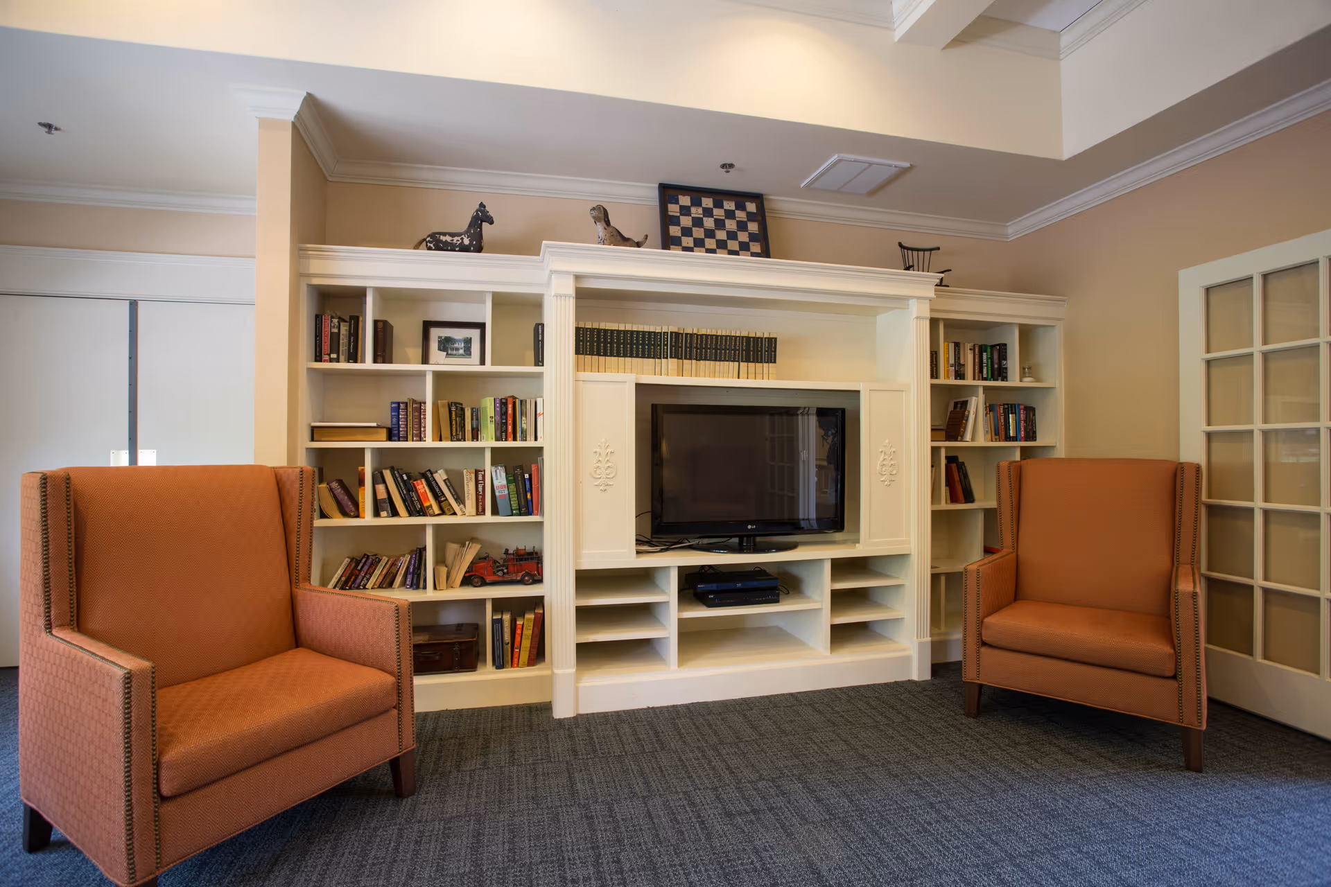 A cozy living room area with two orange upholstered armchairs facing a white built-in entertainment center. The entertainment center has shelves filled with books and decorative items, and a flat-screen TV in the center. The room has beige walls, a carpeted floor, and a glass-paneled door on the right side.