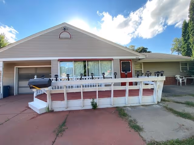 Single-story building with beige siding and a red door, featuring a front porch with a white railing and a ramp. There is a garage door on the left side and a concrete driveway in front. The sky is blue with some clouds.