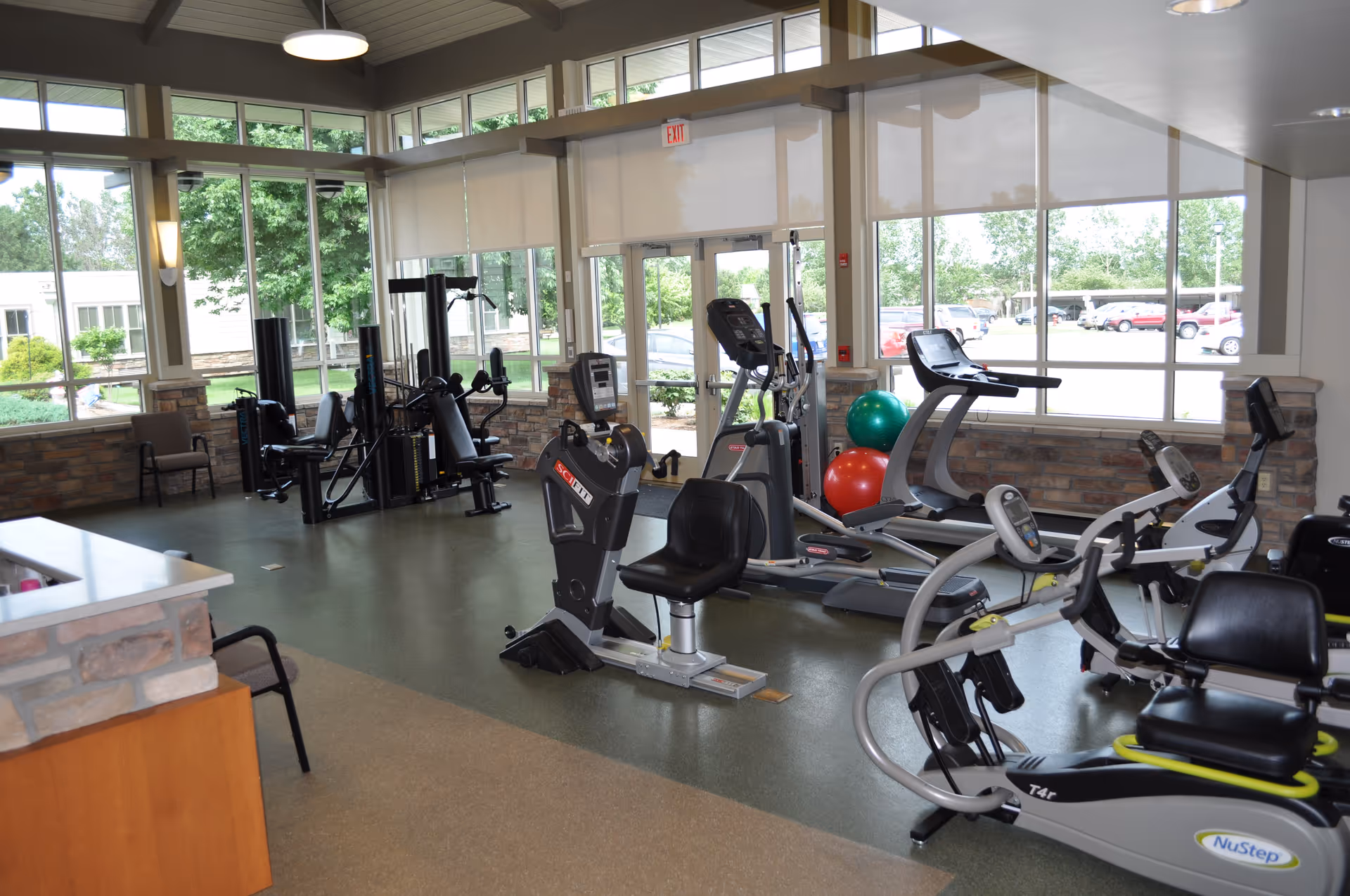 A well-lit fitness room with exercise equipment including recumbent bikes, treadmills, and weight machines. Large windows provide natural light and a view of the outside parking lot and greenery. The room has a clean, modern design with stone accents and a high ceiling.