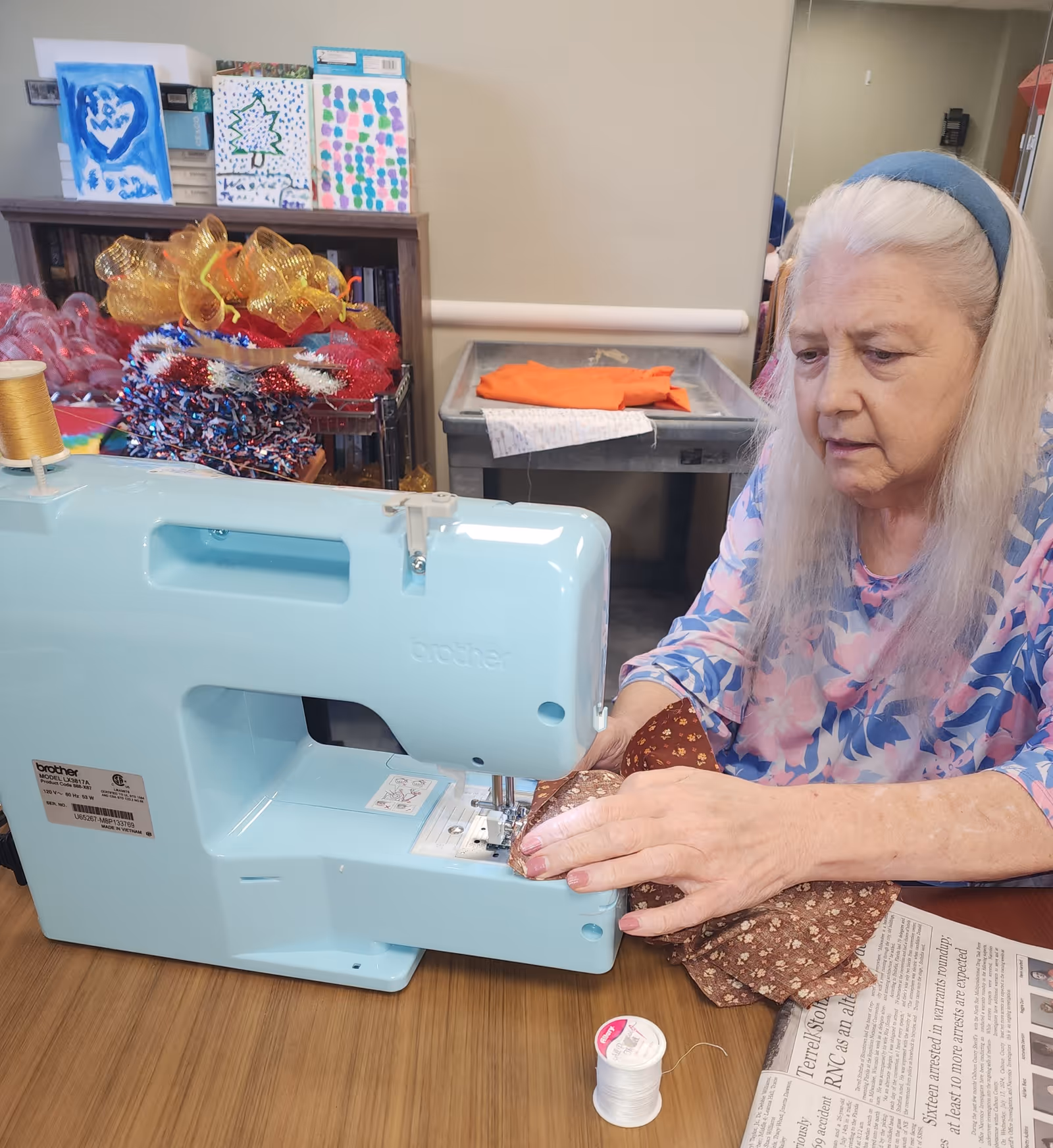 An elderly woman with long white hair and a blue headband is using a light blue Brother sewing machine to sew a piece of brown fabric with a floral pattern. She is seated at a wooden table with a spool of white thread and a newspaper on it. Behind her, there are colorful craft supplies and paintings on a shelf.