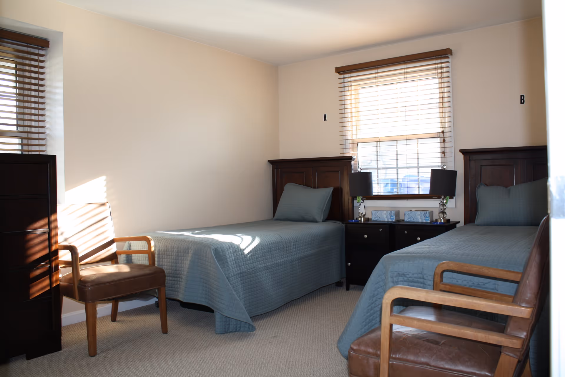 A simple bedroom with two single beds covered in blue quilts, each with a matching pillow. Between the beds is a dark wooden nightstand with two lamps and two tissue boxes. There are two wooden chairs with brown leather seats, one near each bed. The room has beige walls, a window with wooden blinds letting in sunlight, and a dark wooden dresser on the left side.