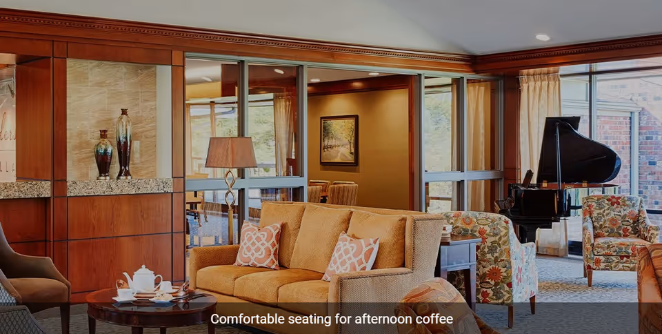 A cozy living room area with a tan sofa adorned with patterned pillows, a round wooden coffee table with a tea set, floral armchairs, a floor lamp, and a grand piano near large windows letting in natural light.