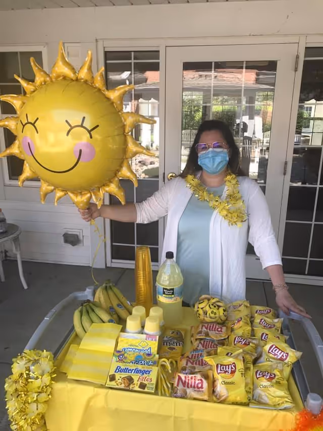 A woman wearing a face mask, glasses, and a yellow flower lei stands behind a table covered with a yellow tablecloth. She is holding a large yellow sun-shaped balloon with a smiling face. The table is filled with yellow-themed snacks including bananas, Lay's Classic potato chips, Nilla wafers, Butterfinger Bites, candy canes, and lemonade. The setting appears to be an outdoor patio area with glass doors in the background.
