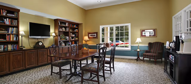 A cozy common room with a wooden dining table and six chairs, built-in wooden bookshelves filled with books and decorative items, a flat-screen TV mounted on the wall, two table lamps, a window showing greenery outside, and a coffee station with a coffee maker and cups on a side cabinet.