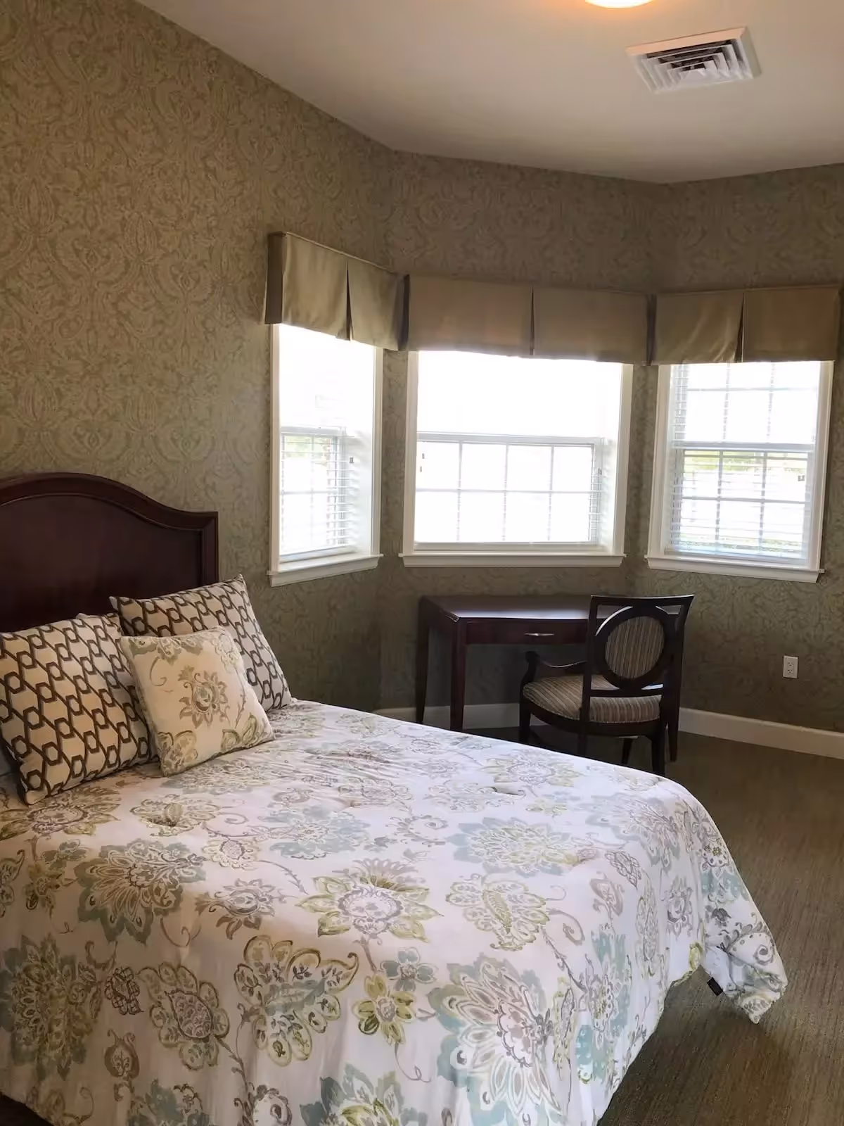 A bedroom with a bed covered in a floral patterned bedspread and three pillows. The room has three windows with beige valances, a wooden desk, and a chair with a cushioned seat. The walls have a subtle patterned wallpaper and the floor is carpeted.
