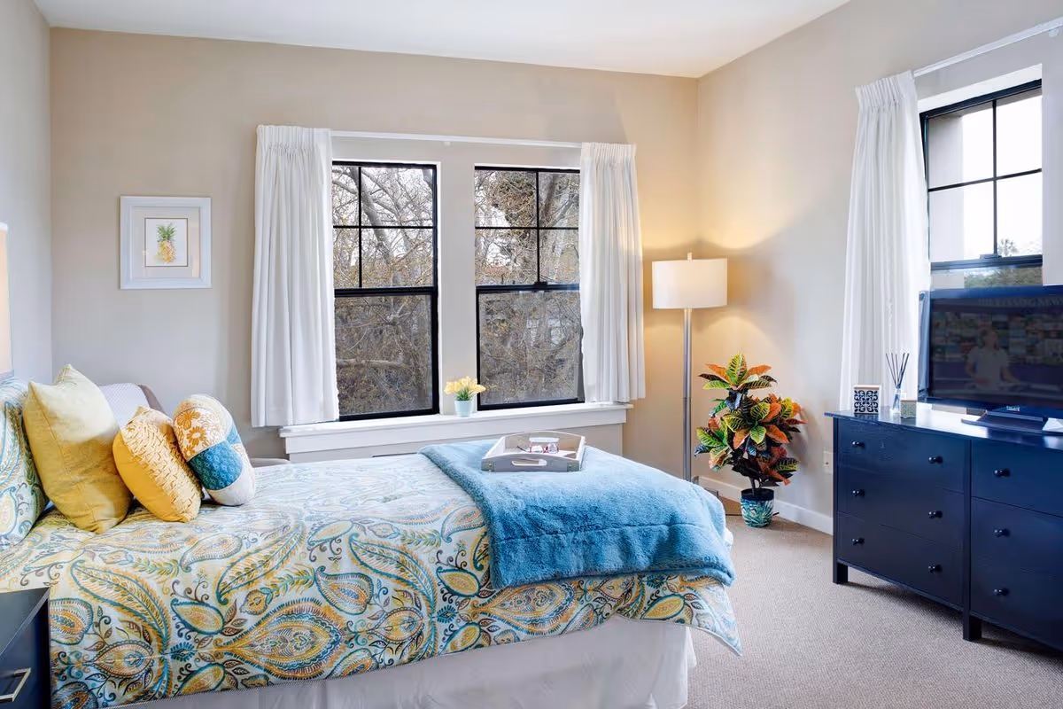 A bright and cozy bedroom with a bed covered in a paisley-patterned bedspread in shades of blue, yellow, and green. The bed has several decorative pillows and a blue throw blanket. There are two large windows with white curtains letting in natural light. A floor lamp and a colorful potted plant are in the corner next to a dark blue dresser with a flat-screen TV on top. A small framed pineapple picture hangs on the wall.