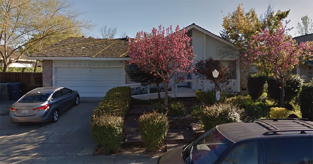 Front exterior of a single-story senior living facility with pink flowering trees and parked cars.