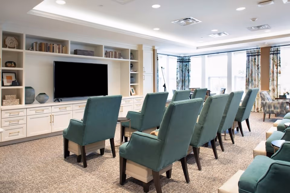 A bright and spacious common area with rows of teal upholstered chairs facing a large flat-screen TV mounted on a white built-in shelving unit filled with books and decorative items. Large windows with patterned curtains allow natural light to fill the room.
