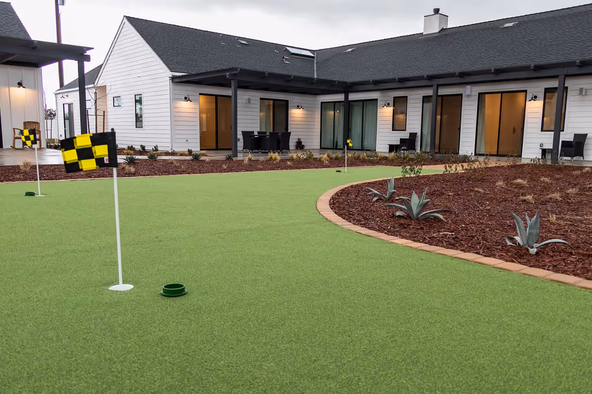 A putting green with small checkered flags in front of a single-story white building with sliding patio doors and outdoor seating.
