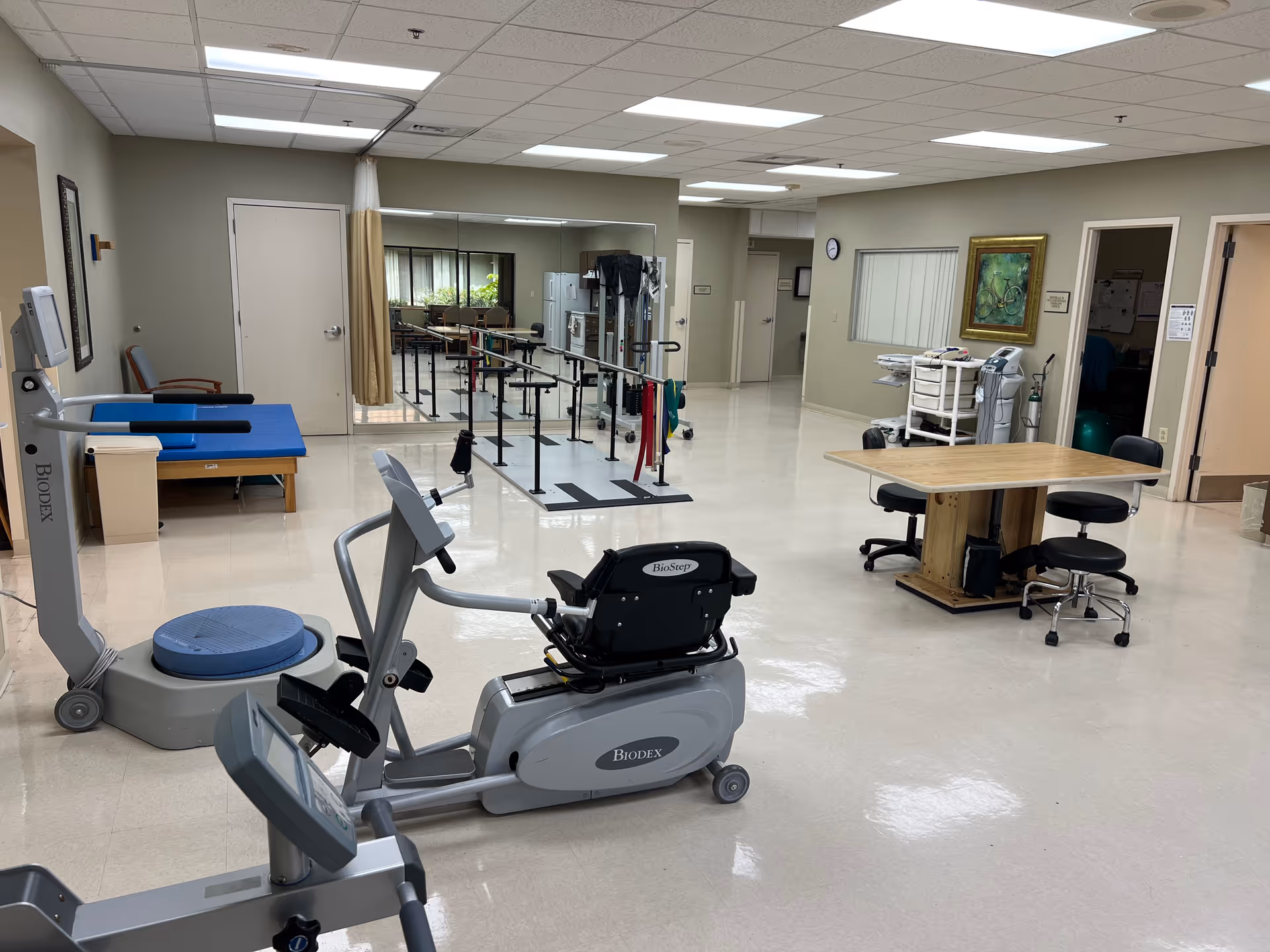 A bright rehabilitation therapy room with exercise machines, parallel bars, a treatment table, and a table with chairs.