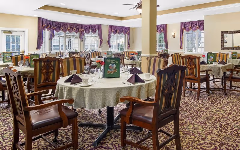 A dining room in a senior living facility with round and rectangular tables covered with white tablecloths. Each table is set with folded purple napkins, cups, glasses, and menus. The room has large windows with purple valances, patterned carpet, and wooden chairs with armrests.