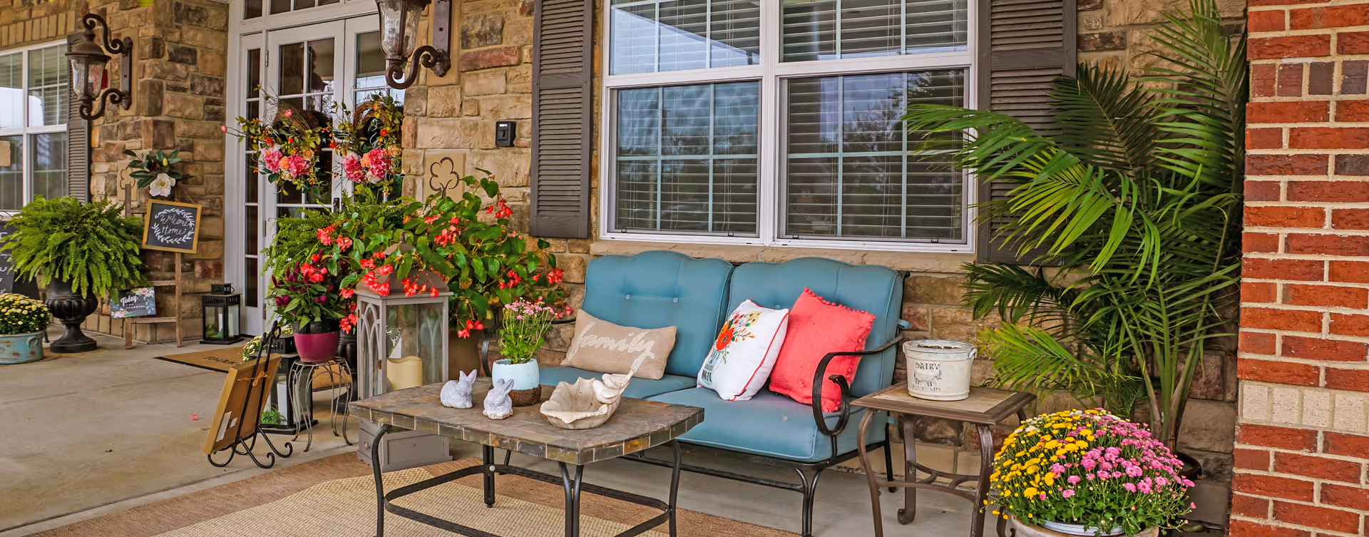 A cozy outdoor seating area at the entrance of a building with a blue cushioned bench adorned with colorful pillows, a coffee table with decorative items including small rabbit statues and a shell, surrounded by various potted plants and flowers. The background features stone and brick walls, large windows with shutters, and double glass doors with floral wreaths. There are also lanterns and welcome signs near the entrance.