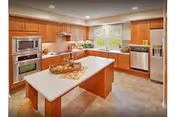 A spacious kitchen with wooden cabinets, a central island with a basket of fruit and a cutting board, stainless steel appliances including a microwave, oven, dishwasher, and refrigerator, and a window above the sink letting in natural light.