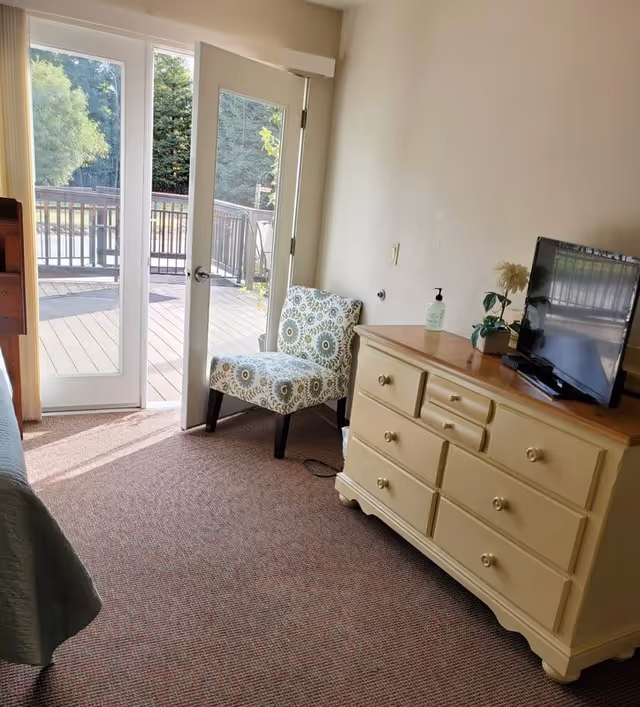 A cozy senior living room area with a patterned upholstered chair next to a glass door leading to an outdoor deck. A cream-colored dresser with multiple drawers holds a flat-screen TV, a small potted plant, a flower, and a bottle of hand sanitizer. The room is carpeted and well-lit with natural light coming through the door.