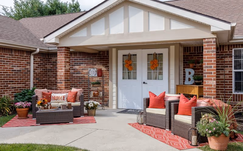 Outdoor seating area in front of a brick building entrance with white double doors decorated with orange wreaths. The area features two sets of wicker chairs and a loveseat with red and beige cushions, red patterned rugs, potted plants, and decorative items including a 'HOME' pillow and a large letter 'B'.