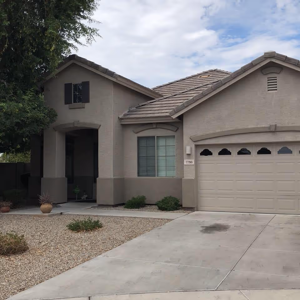 Exterior view of a single-story residential building with a beige stucco finish, a tiled roof, a two-car garage with windows, a driveway, and a small landscaped front yard with gravel and some plants.