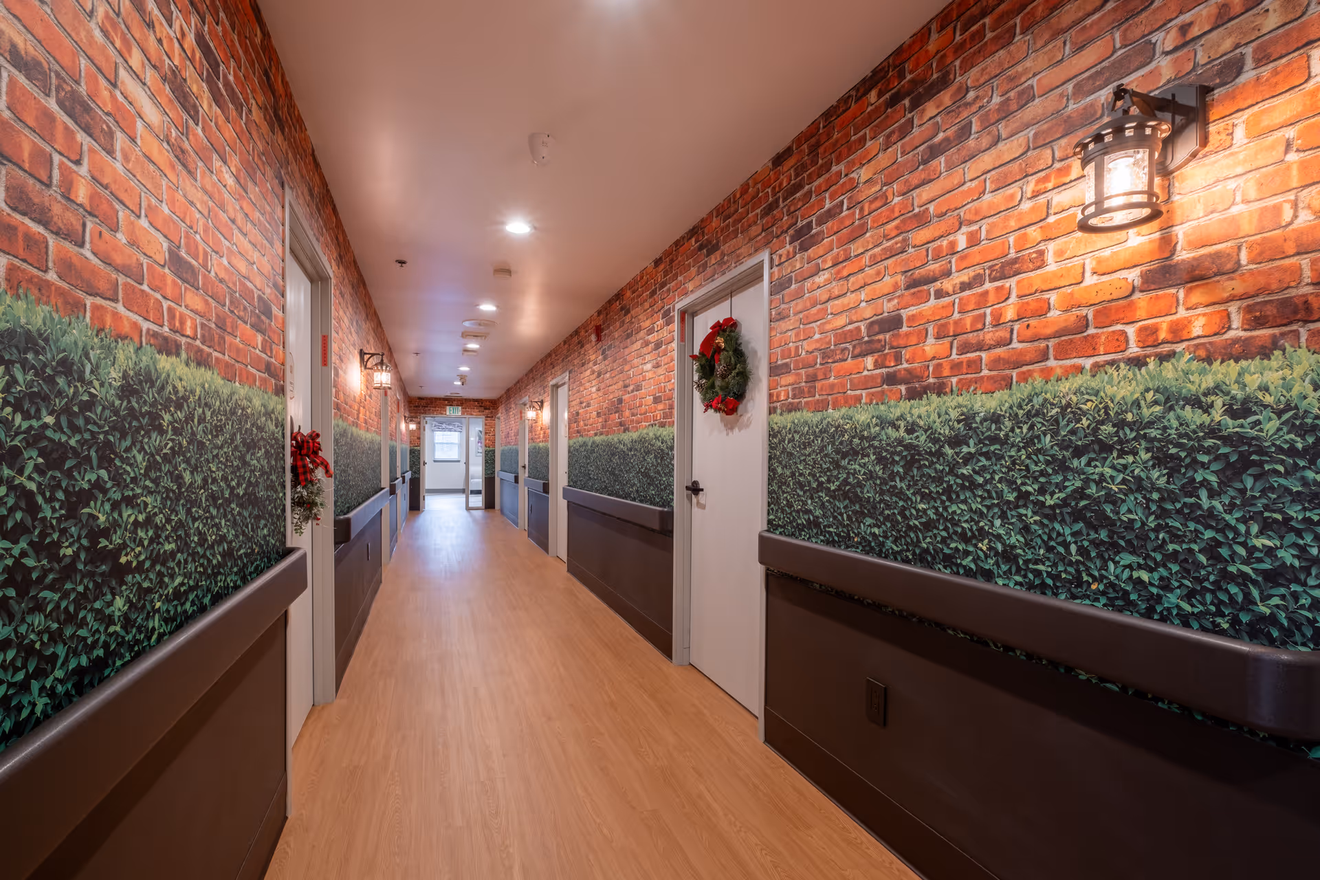 A long indoor hallway with wood flooring and walls decorated with a mural of brick and green shrubbery. Several white doors line the hallway, each adorned with a holiday wreath. Wall-mounted lantern-style lights provide warm lighting along the corridor.