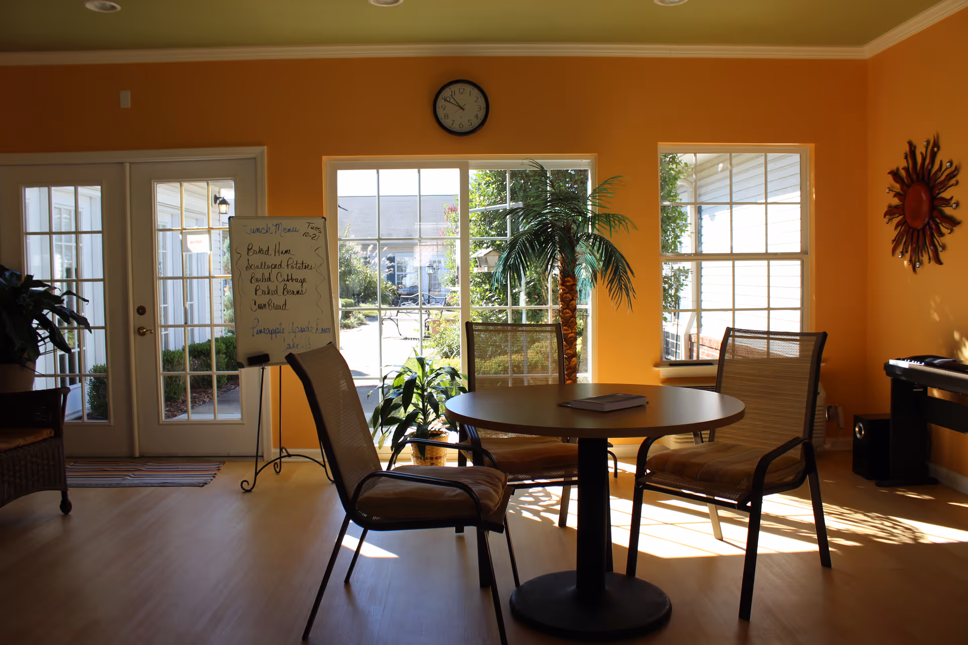 A bright room with orange walls featuring a round table surrounded by three chairs. Large windows and glass doors let in natural light, showing an outdoor garden area. A clock is mounted above the central window, and a whiteboard with a lunch menu is positioned near the doors. There are several plants, including a tall palm, and a decorative sun wall hanging on the right wall.
