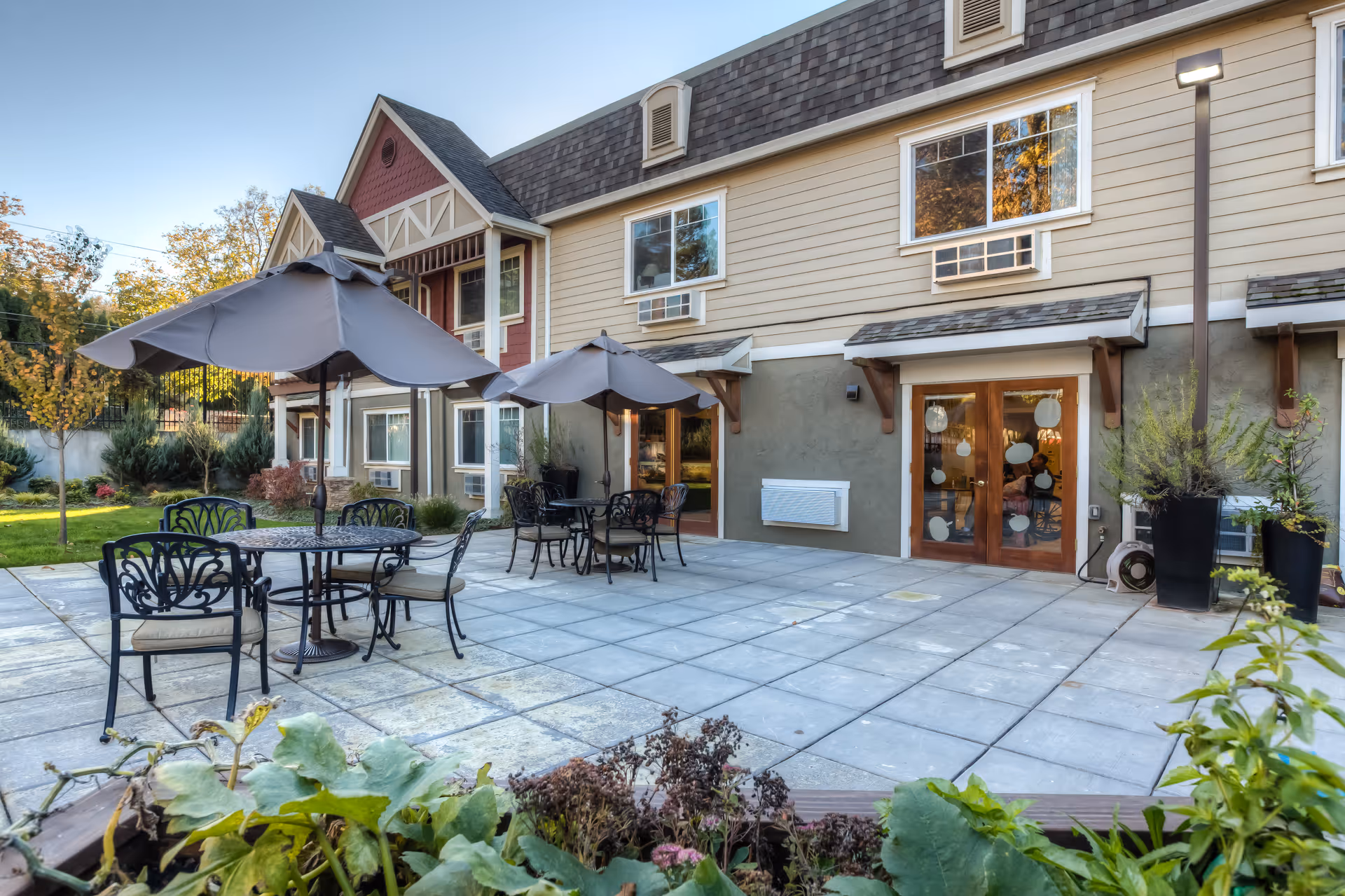 Outdoor patio with tables, umbrellas, and chairs in front of a two-story building entrance.