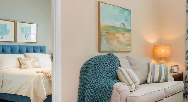 A cozy living room area with a light gray sofa adorned with striped pillows and a teal knitted throw blanket. A table lamp with a beige shade sits on a side table next to the sofa. On the wall above the sofa is a framed painting of a landscape with a small house. Through an open doorway, a bedroom is visible with a blue upholstered headboard, white bedding, and two framed leaf prints on the wall.