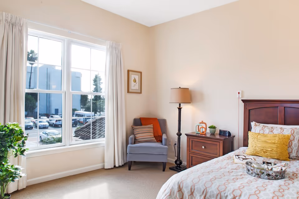 A bright bedroom with a large window showing a parking lot outside. Inside, there is a gray armchair with a striped pillow and an orange throw blanket, a wooden nightstand with a lamp, decorative items, and an alarm clock. The bed has a patterned bedspread with a yellow pillow and a tray with glasses and a book.