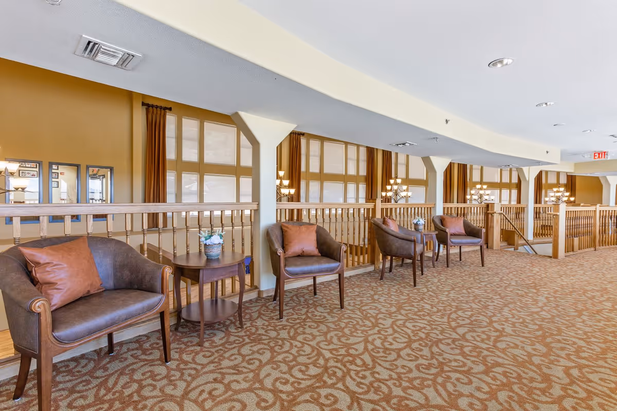 A spacious interior hallway area with patterned carpet, featuring a row of brown leather armchairs each paired with a small wooden side table holding a flower arrangement. The area is well-lit with large windows covered by shades and brown curtains, and decorative light fixtures are visible in the background.
