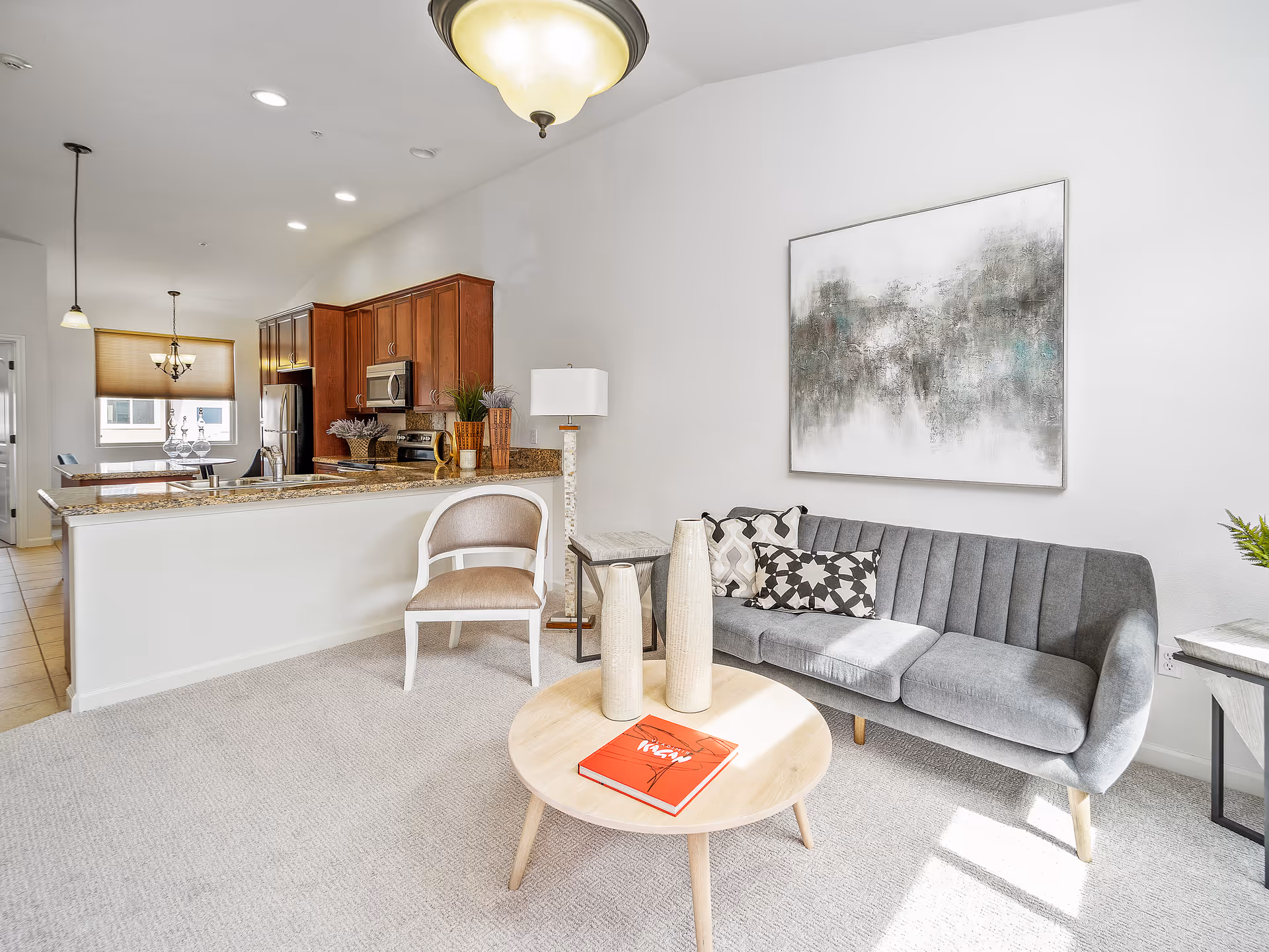 Bright open-plan living room showing a gray sofa, round coffee table with vases, and a granite-topped kitchen counter in the background.