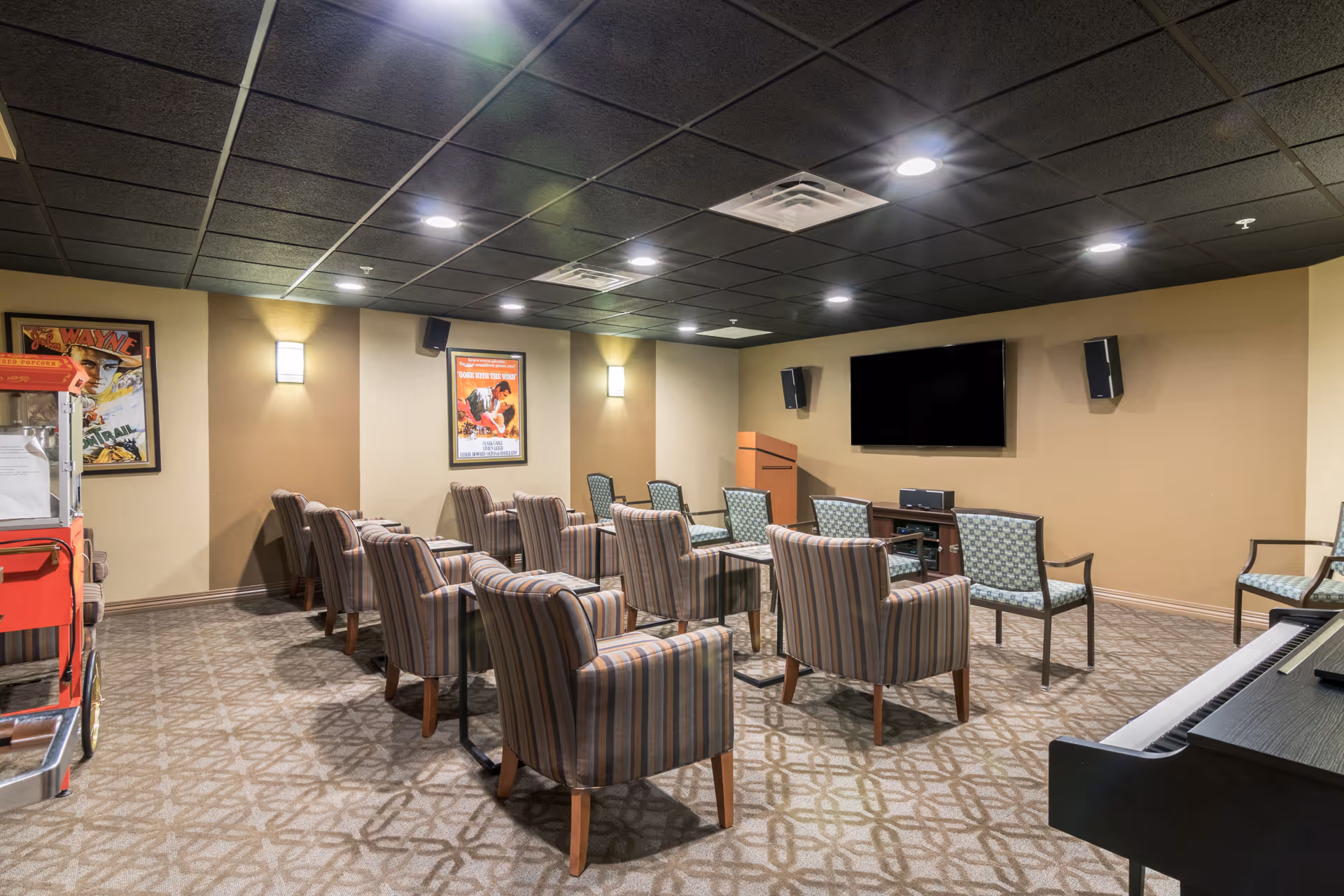 A cozy media room with multiple striped and patterned armchairs arranged in rows facing a large flat-screen TV mounted on a beige wall. The room has a patterned carpet, a popcorn machine on the left, wall-mounted speakers, and framed movie posters on the walls. The ceiling is black with recessed lighting.